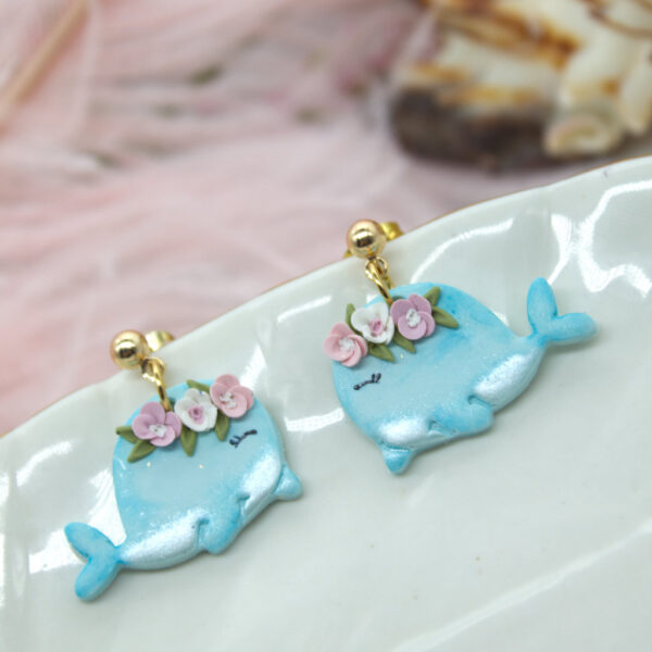 Handmade polymer clay earrings in the shape of cute blue whales, adorned with small flowers on their heads, perfect for a summer-themed outfit