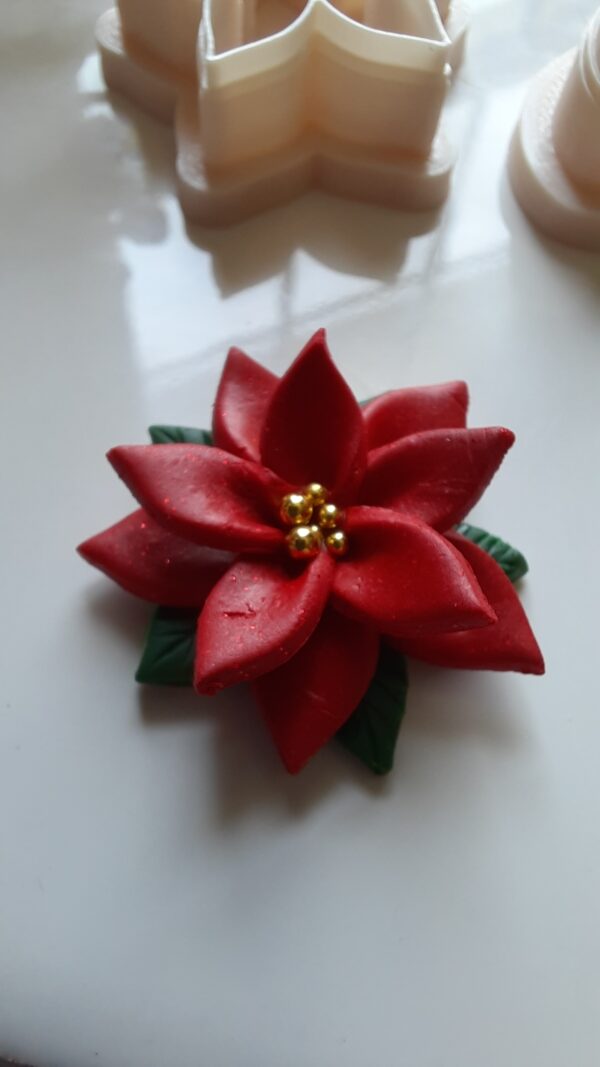 A close-up photo of a poinsettia flower made of polymer clay with six petals and a cluster of small yellow beads in the center, representing the stamen. The flower is placed on a white surface, and there are polymer clay cutters for this poinsettia in background