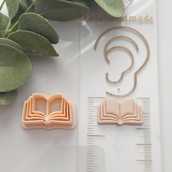 Polymer Clay Cutter in the shape of an open book, alongside a crafted polymer clay earring, surrounded by leaves